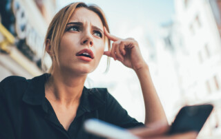 From below of confused blond haired female entrepreneur in black shirt looking away in though while solving problems with partner on mobile phone at city street